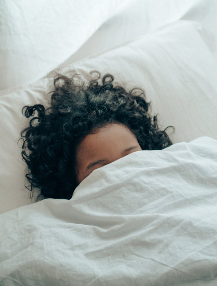 A woman with dark curly hair lies in bed with the blanket covering her face