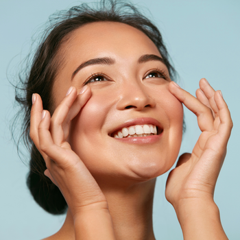 A lady standing out in the sun smiling while touching her face.