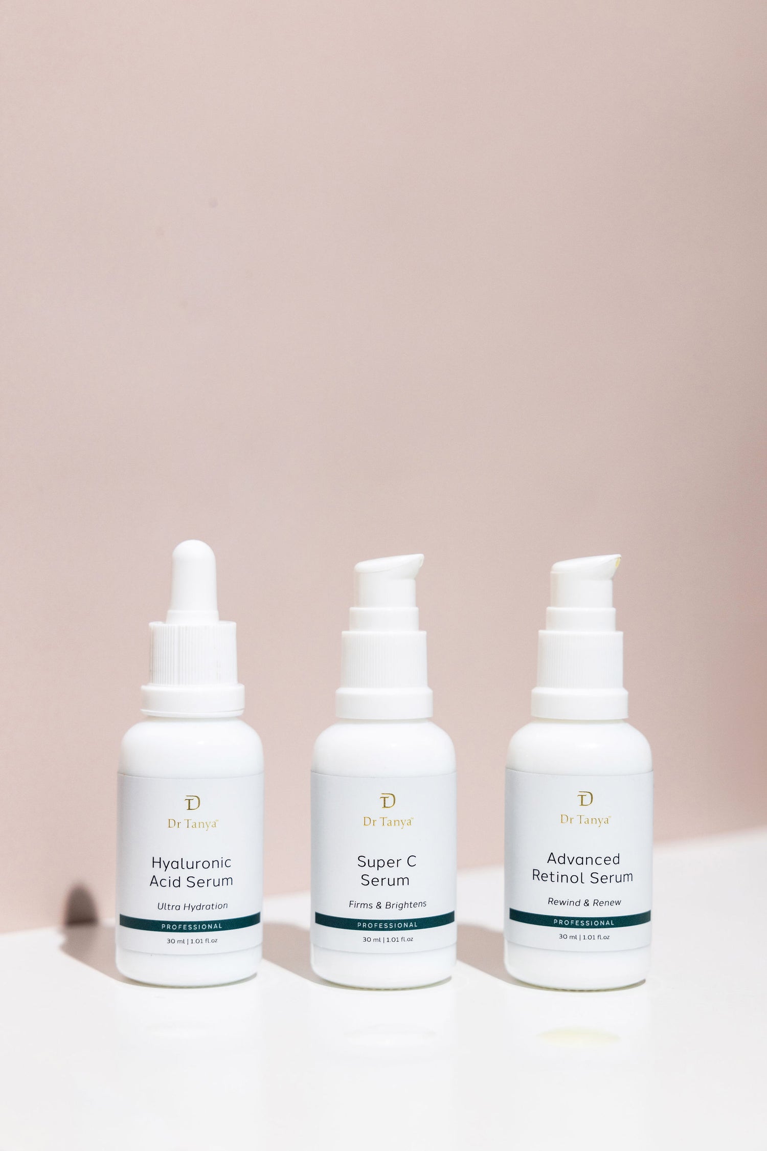 Three white bottles of face serum in front of a blush pink wall